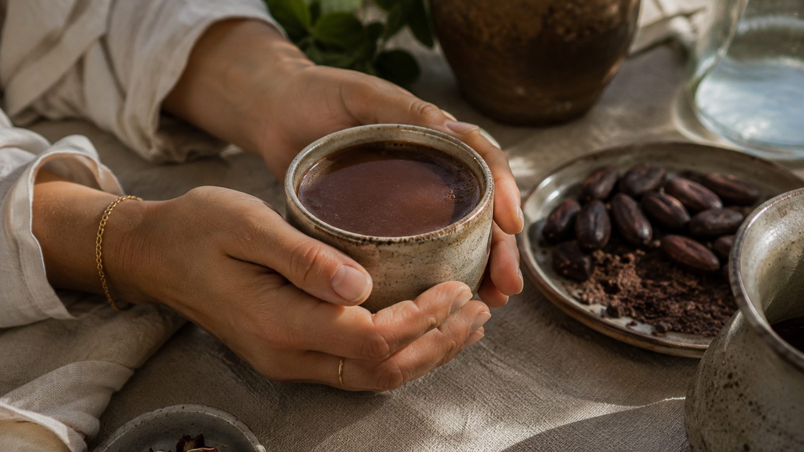 Hands cupping a ceramic bowl of ceremonial cacao on a linen table, cacao beans on a plate beside it
