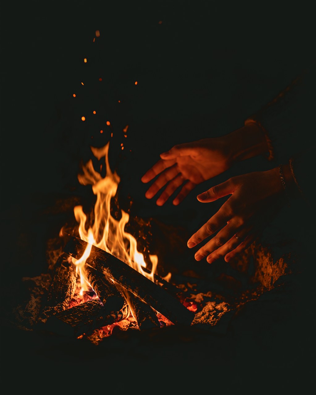 Two hands held open to a low fire at night, sparks rising into the dark